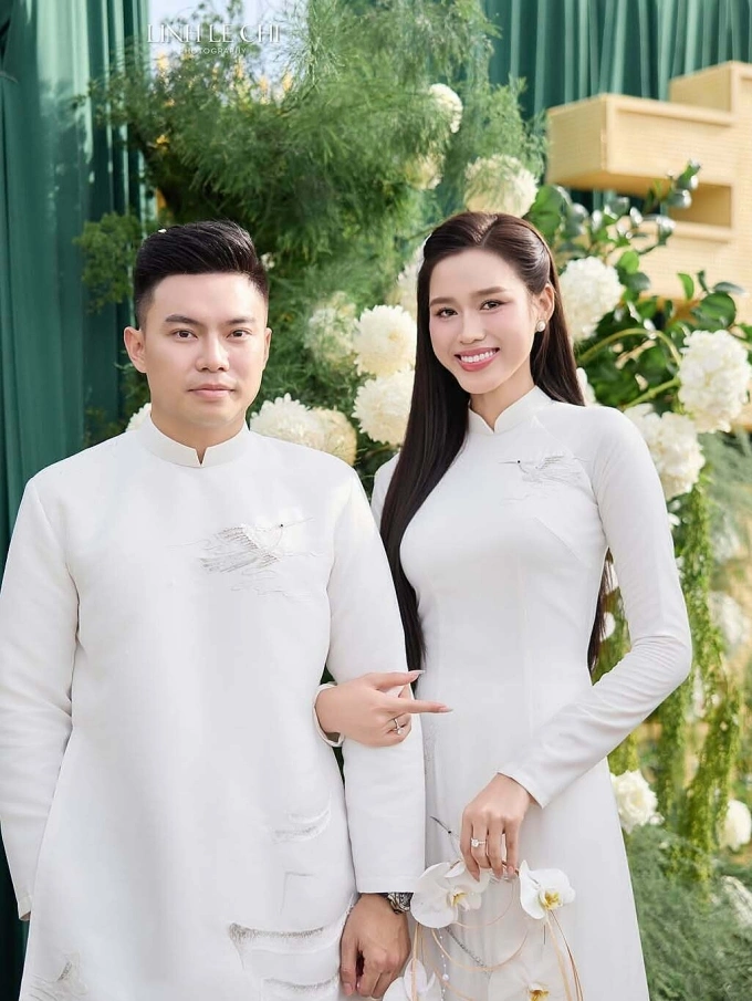 Vợ chồng Đỗ Thị Hà diện áo trắng thêu họa tiết cánh chim. Hoa hậu cầm vòng hoa lan thay cho bó hoa cưới thông thường, tạo sự khác biệt.
